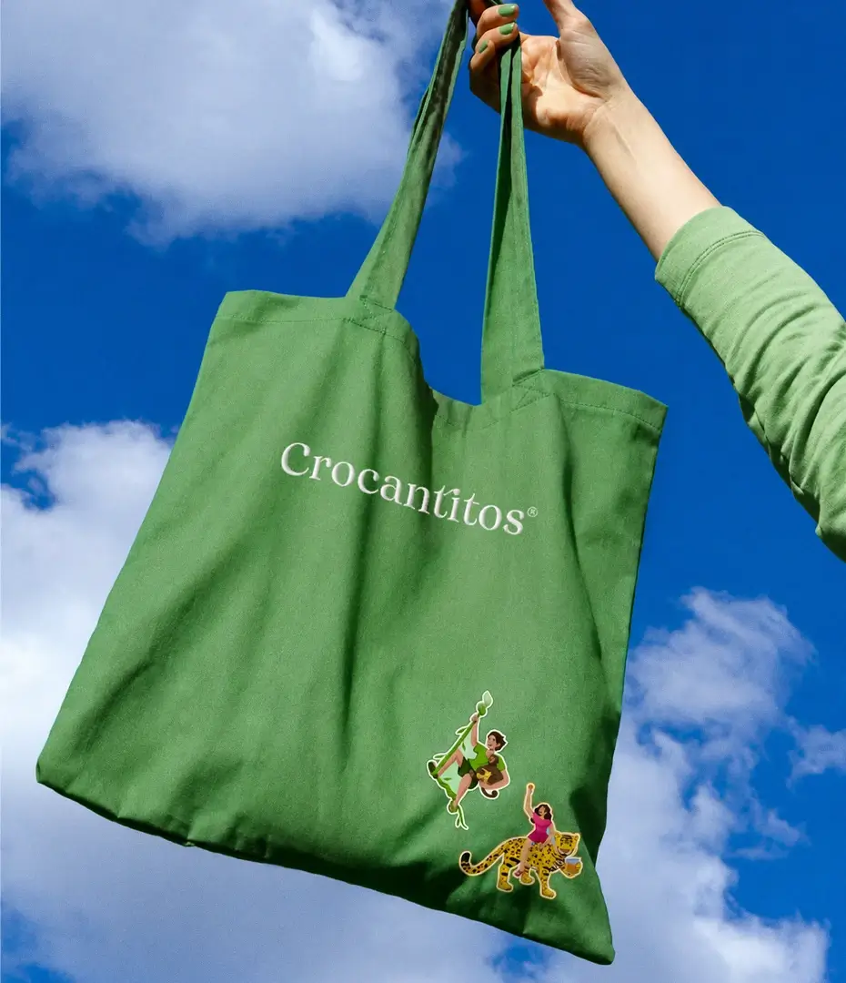 Bolsa de tela tote bag ecológica de color verde con el logotipo de Crocantitos e ilustraciones promocionales sostenida en alto contra un cielo azul por BSM.